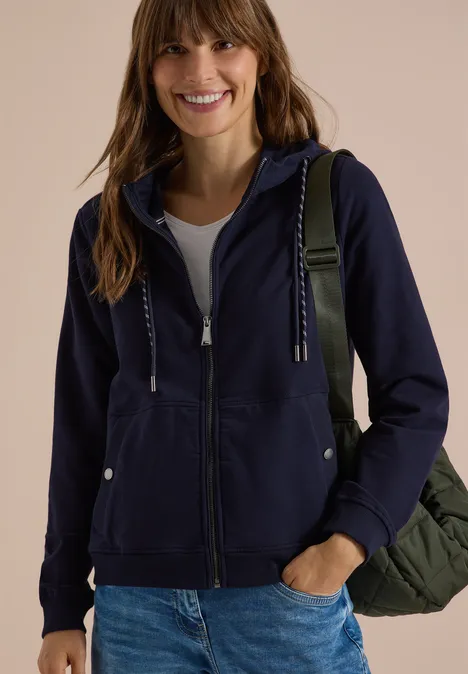 Junge Frau in dunkelblauer Kapuzenjacke und Jeans, lächelnd, mit einer Tasche über der Schulter.