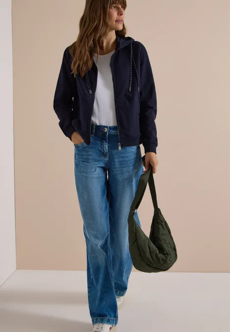 Frau in dunkelblauer Jacke, weißem Top und weiter Jeans, die eine grüne Umhängetasche trägt.