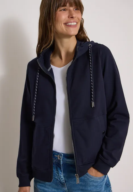 Frau trägt dunkelblaue Kapuzenjacke, weißes Shirt und Jeans, lächelt und hat die Hände in den Taschen.