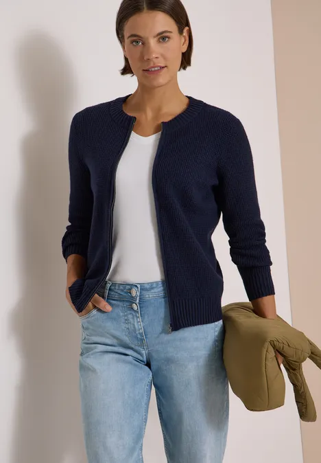 Femme en gilet bleu marine, top blanc et jean, tenant une veste kaki négligemment sur le bras.