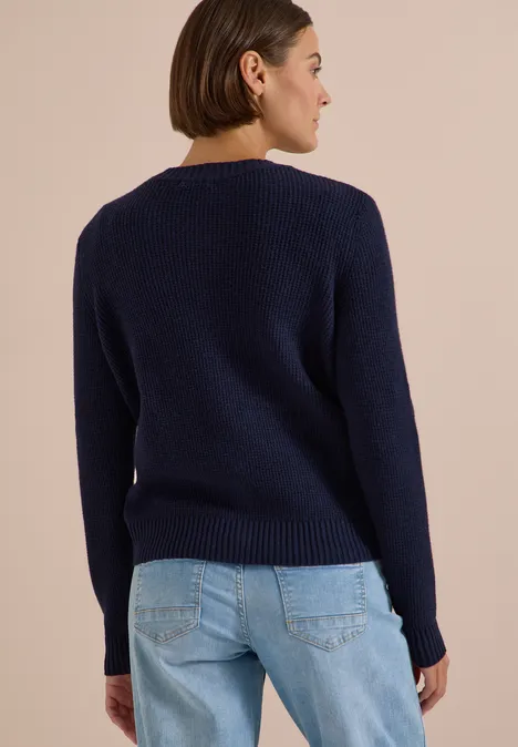 Femme vue de dos portant un pull bleu foncé et un jean bleu clair sur fond clair.
