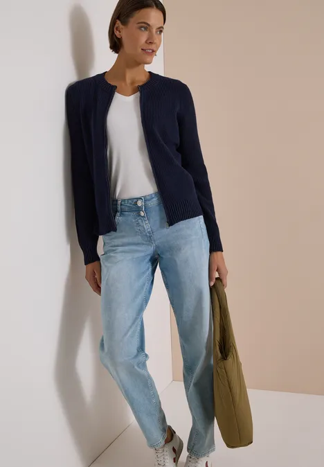 Femme en t-shirt blanc, jean clair, gilet bleu marine, sac vert devant un mur bicolore.