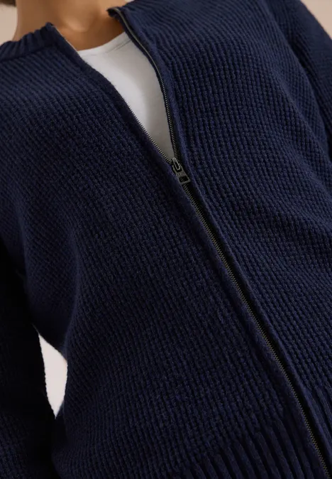 Gilet zippé bleu marine en maille épaisse, ouvert sur un haut blanc visible en dessous.