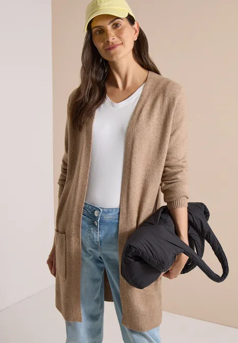 Vrouw leunt tegen muur, draagt beige vest, wit topje, spijkerbroek en gele pet, houdt een zwarte tas vast.