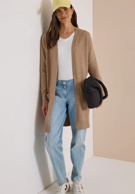 Vrouw in lange beige cardigan, wit T-shirt, lichtblauwe jeans, witte sneakers, houdt gele pet en zwarte jas vast