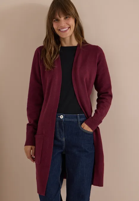 Cardigan long à la coupe ample red wine Femme souriante portant un gilet bordeaux, un t-shirt noir et un jean bleu sur fond neutre.
