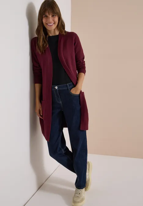 Cardigan long à la coupe ample red wine Une femme souriante appuyée contre un mur, portant un cardigan bordeaux, un pull, un jean et des chaussures claires.