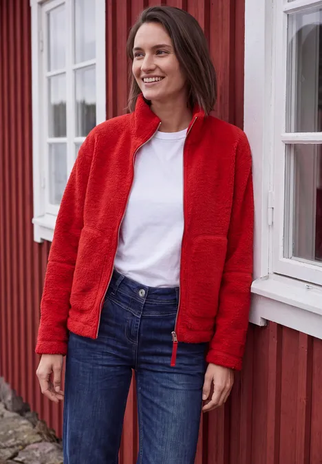 Teddyjacke mit Stehkragen und Zipper flame red Lächelnde Frau in roter Fleecejacke, weißem Shirt und Jeans lehnt vor einem roten Haus mit weißen Fensterrahmen.