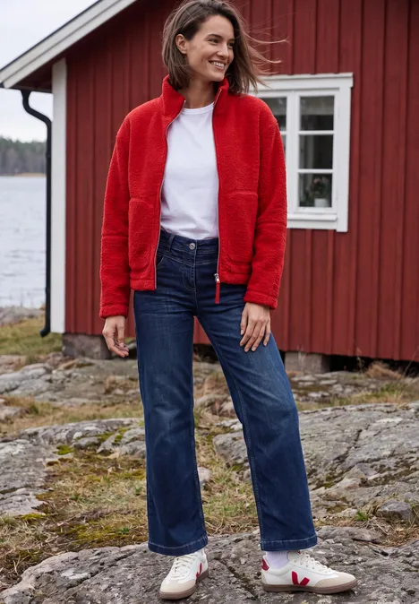 Teddyjacke mit Stehkragen und Zipper flame red Frau in roter Jacke, weißem Shirt und Jeans steht vor einem roten Holzhaus am See auf felsigem Untergrund.