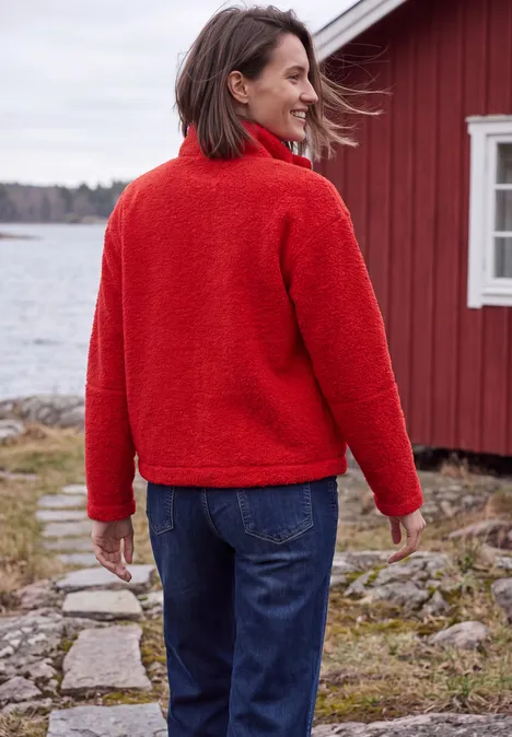 Teddyjacke mit Stehkragen und Zipper flame red Frau in roter Fleecejacke und Jeans steht am Seeufer vor einem roten Haus.