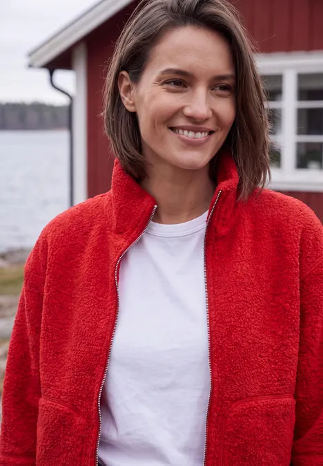 Teddyjacke mit Stehkragen und Zipper flame red Eine lächelnde Frau in roter Fleecejacke und weißem T-Shirt steht vor einem roten Haus am See.