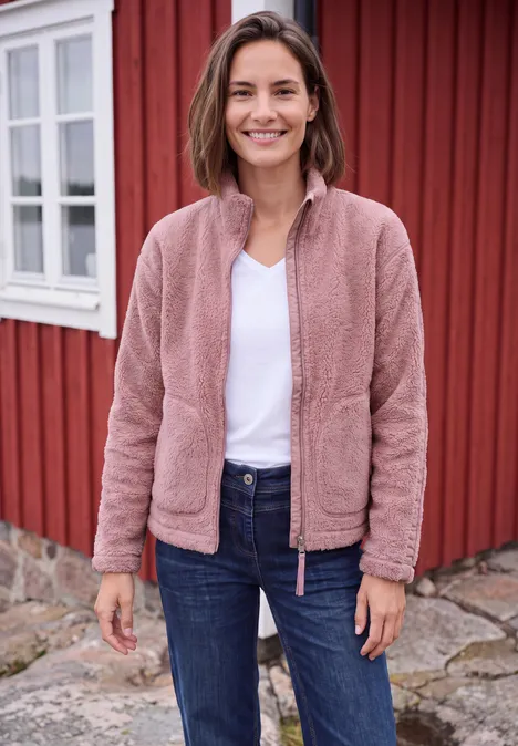 Teddyjacke mit Stehkragen und Zipper velvet rose Eine lächelnde Frau in rosa Fleecejacke, weißem T-Shirt und Jeans vor einem roten Holzhaus.
