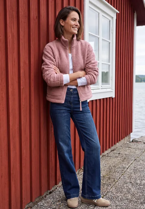 Teddyjacke mit Stehkragen und Zipper velvet rose Eine lächelnde Frau in rosa Jacke und Jeans lehnt entspannt an einem roten Holzhaus am Wasser.