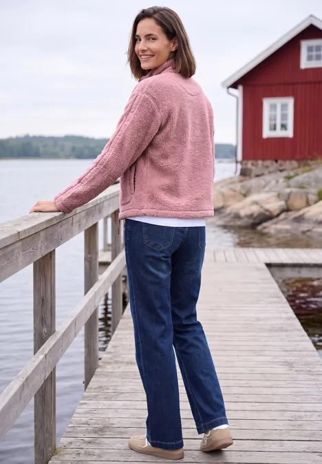 Teddyjacke mit Stehkragen und Zipper velvet rose Frau in rosa Jacke und Jeans lehnt an Geländer auf einem Holzsteg an einem roten Haus am Wasser.
