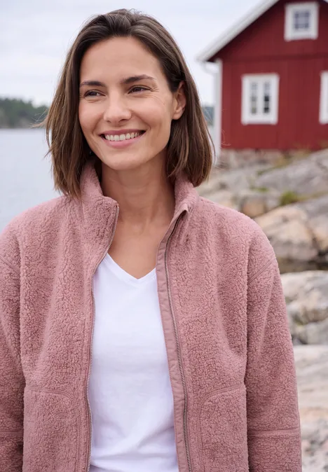 Teddyjacke mit Stehkragen und Zipper velvet rose Eine lächelnde Frau in rosa Fleecejacke vor einem roten Haus am Wasser.
