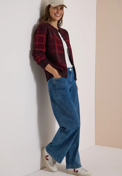 Federgarn Cardigan mit Rundhals red wine Frau lehnt lächelnd an Wand, trägt karierte Strickjacke, weißes Shirt, weite Jeans und Sneaker.