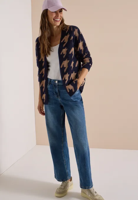 Femme en jean droit, top blanc, gilet pied-de-poule et casquette, souriante devant un fond neutre.