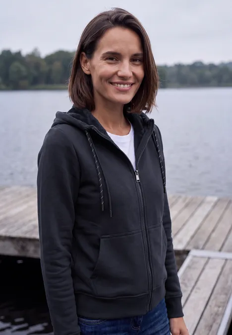 Eine junge Frau in Kapuzenjacke steht auf einem Steg am See und lächelt in die Kamera.