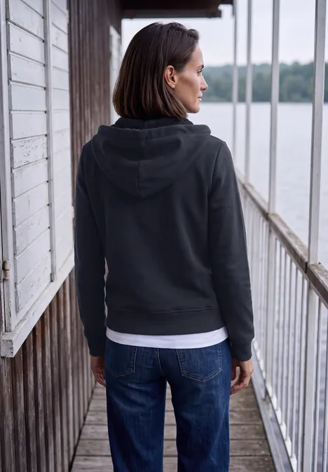 Frau in schwarzer Kapuzenjacke und Jeans steht auf hölzernem Steg und blickt aufs Wasser.