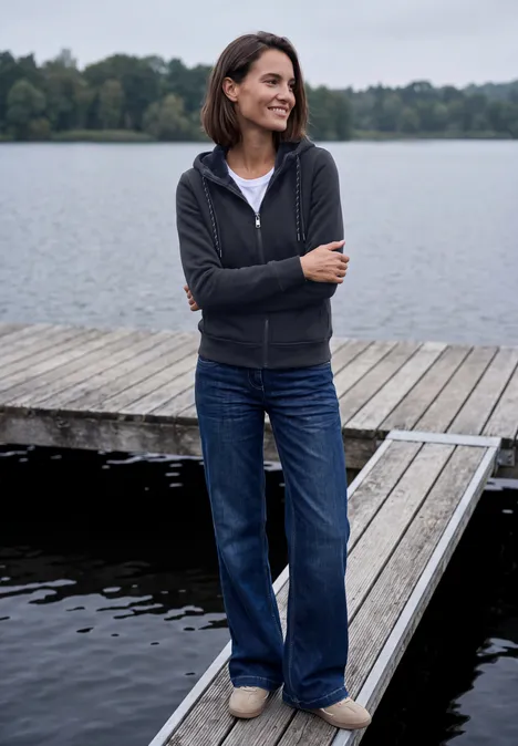 Eine Frau steht lächelnd am Ende eines Holzstegs am See, trägt Jeans und eine dunkle Kapuzenjacke.