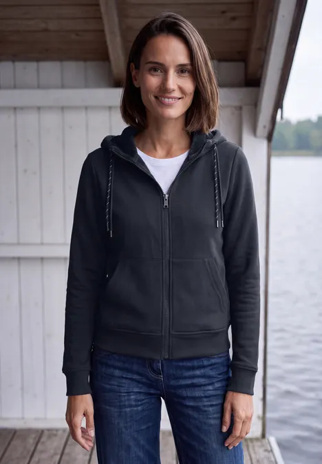Eine Frau steht lächelnd vor einer Seehütte, trägt Kapuzenjacke, T-Shirt und Jeans.