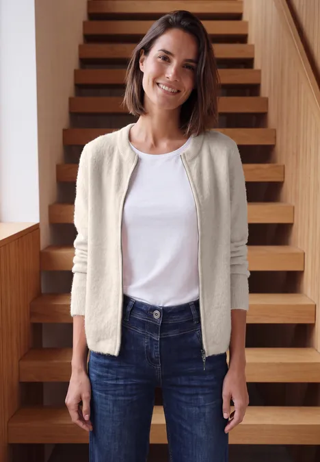 Een glimlachende vrouw in een wit T-shirt, licht beige vest en spijkerbroek staat voor een houten trap.