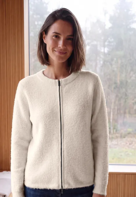 Een vrouw draagt een crèmekleurige teddyfleecejas met rits, staand voor een raam met boszicht.