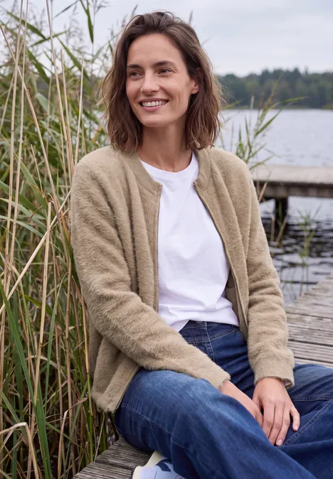 Federgarn Cardigan mit Rundhals creamy mocha melange Frau in beige Strickjacke und Jeans sitzt lächelnd am Seeufer neben Schilf auf einem Steg.