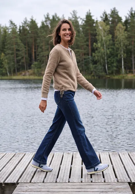 Federgarn Cardigan mit Rundhals creamy mocha melange Frau spaziert lächelnd auf einem Steg am See, umgeben von Bäumen.