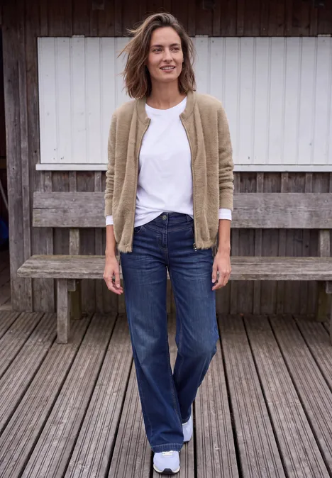 Federgarn Cardigan mit Rundhals creamy mocha melange Eine Frau in lässiger Kleidung steht vor einer Holzbank und lächelt in die Kamera.