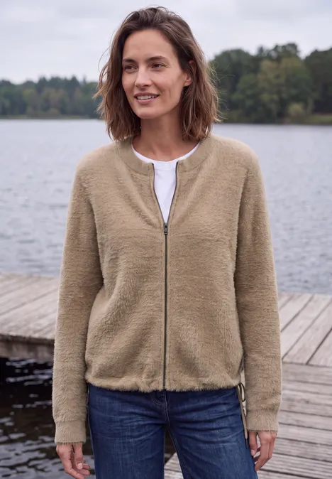 Federgarn Cardigan mit Rundhals creamy mocha melange Frau in beiger Jacke steht entspannt an einem Holzsteg vor einem ruhigen See.