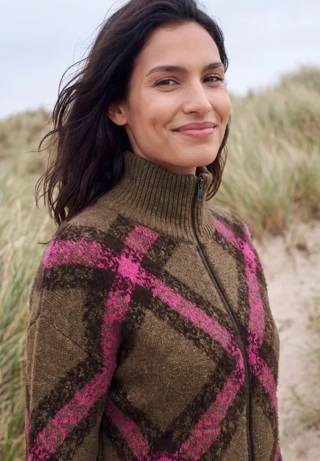 Frau am Strand in einem braunen Wollpullover mit pinkem Karomuster und Reißverschluss.