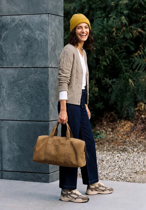 Cardigan mit V-Neck und Zopfmuster toffee cream melange Eine lächelnde Frau in gelber Mütze trägt eine Tasche und steht vor einer Mauer mit grünem Hintergrund.