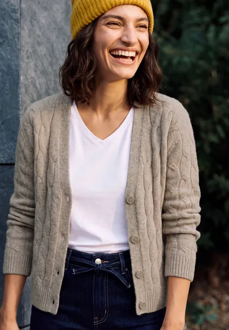 Cardigan mit V-Neck und Zopfmuster toffee cream melange Eine Frau mit gelber Mütze, weißem T-Shirt und grauer Strickjacke lächelt im Freien vor Grün.