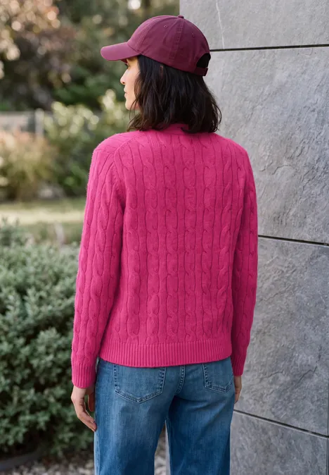Frau mit pinkem Strickpullover, blauen Jeans und roter Cap steht rückwärts vor einer grauen Wand im Freien.