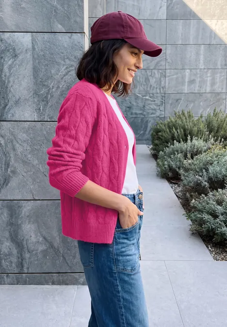 Eine junge Frau im pinken Cardigan, Jeans und roter Mütze steht seitlich lächelnd vor einer Steinwand.