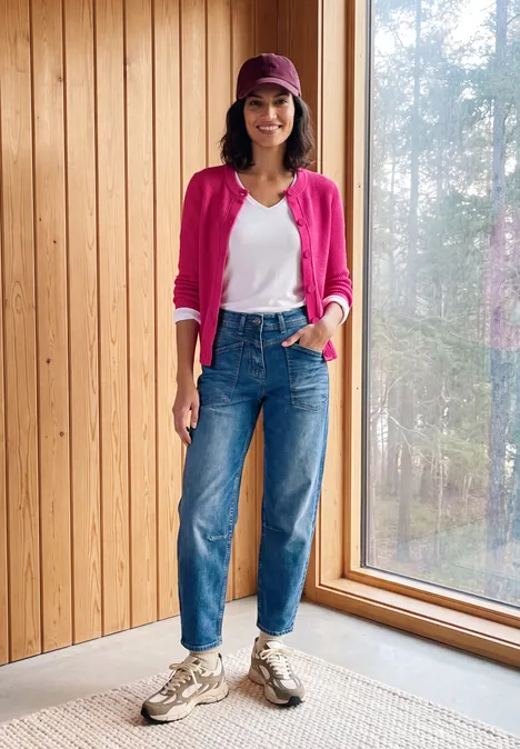 Frau in rosa Cardigan, weißem Shirt, Jeans und Cap steht lächelnd am Fenster vor Holzwand.