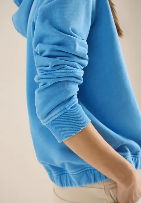 Personne en sweat bleu et pantalon beige, vue de côté sur fond clair.