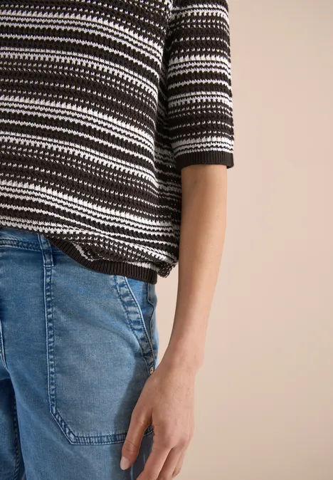 Nahaufnahme eines Oberkörpers in gestreiftem Pullover und Jeans vor beigem Hintergrund.