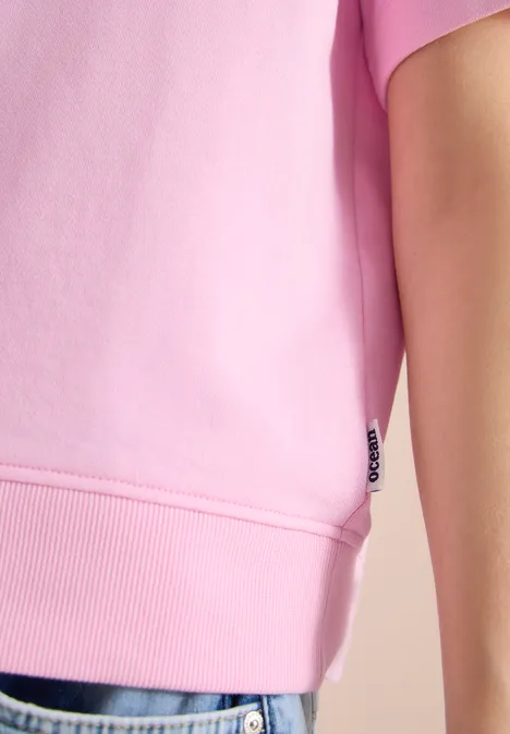 Person trägt hellrosa Kurzarmshirt mit Bündchen und Jeans mit ausgefranstem Saum.