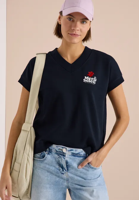 Person trägt schwarzes V-Neck T-Shirt mit kleinem weiß-rotem Logo, Jeans und beige Umhängetasche vor hellem Hintergrund.