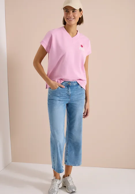 Frau trägt pinkes T-Shirt, hellblaue, weite Cropped-Jeans und weiße Sneaker vor beigem Hintergrund.