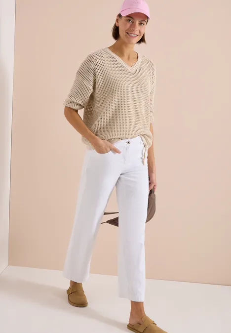 Eine Person trägt einen beigen V-Ausschnittpullover, weiße Jeans und braune Loafers.