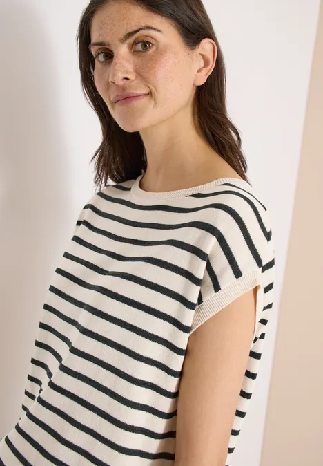 Frau trägt kurzärmliges weißes Shirt mit schwarzen Querstreifen, vor hellem Hintergrund, Gesicht verschwommen.