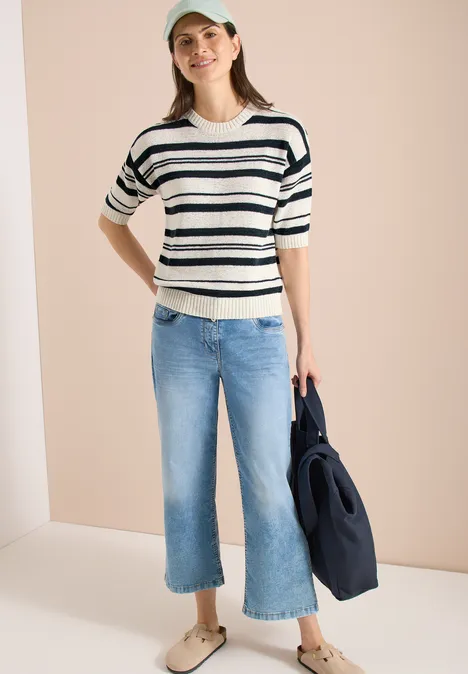 Frau in gestreiftem Pullover, weitem Jeans-Culotte und dunkler Tasche vor hellem Hintergrund.