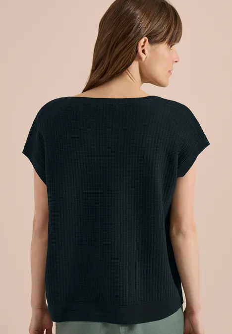 Vrouw van achteren in een zwart ribgebreid shirt met korte mouwen voor een beige achtergrond.