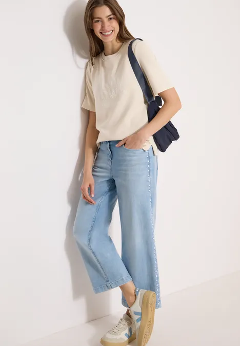 Person in weißem Shirt, heller Jeans-Culotte, Sneakers und navy Schultertasche lehnt an Wand.