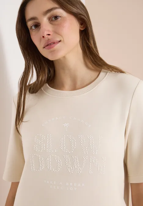 Eine Frau trägt ein cremefarbenes T-Shirt mit auffälligem „SLOW DOWN“-Schriftzug aus Nieten.