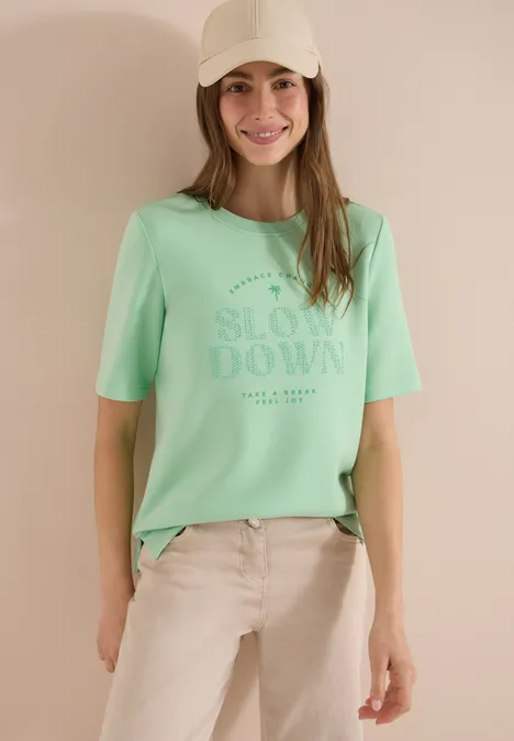 Frau trägt mintgrünes T-Shirt mit Aufdruck 'SLOW DOWN', vor neutralem Hintergrund.