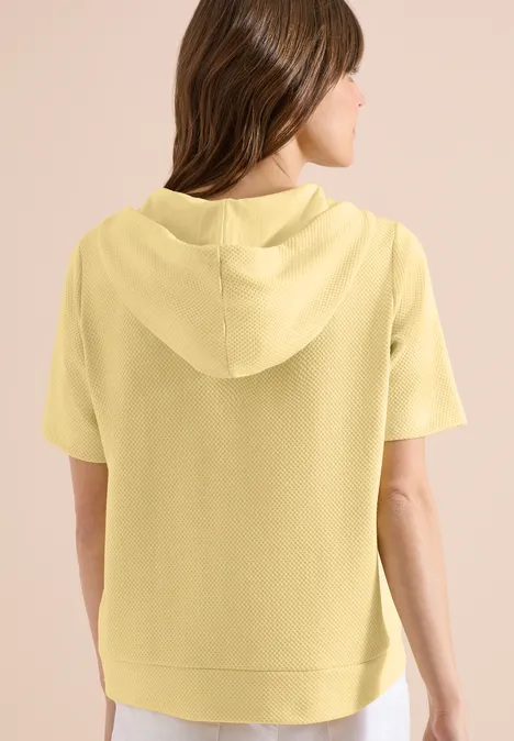 Frau von hinten in einem hellgelben Kurzarm-Hoodie vor hellem Hintergrund.